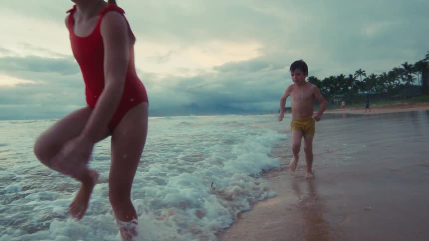 Three joyful children run and splash in the ocean waves along the sandy shore at dusk in slow motion