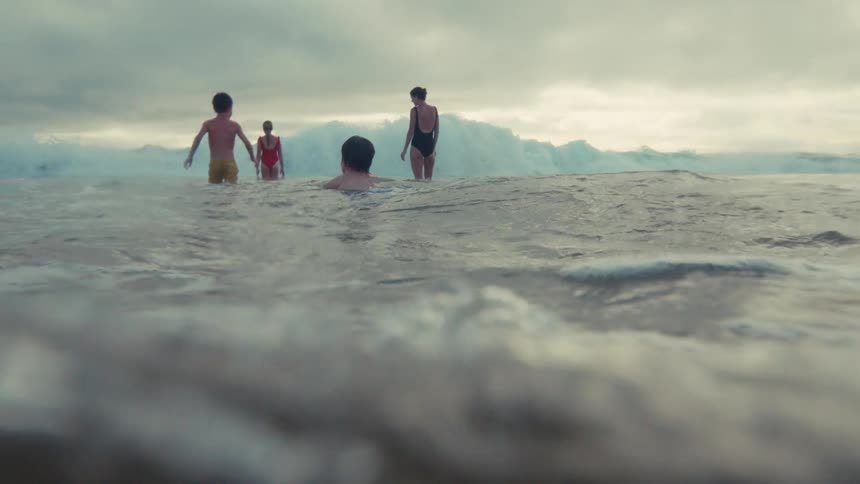 An immersive slow-motion view from the water's surface shows a family waiting for waves on a moody, overcast day