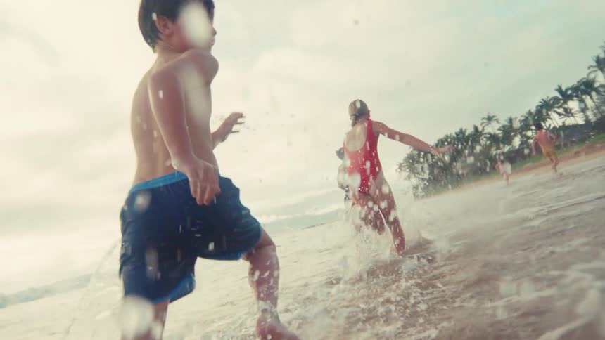 Happy children run and splash through the shallow ocean waves in slow motion on a tropical beach