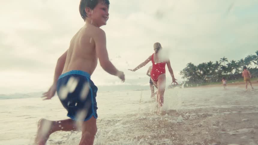 Happy children run and splash through the shallow ocean waves in slow motion on a tropical beach