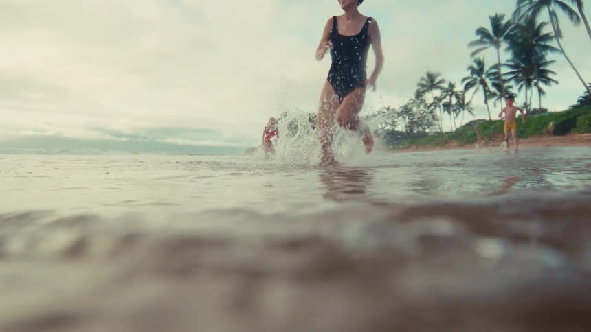A slow motion, low angle shot of a woman and children joyfully running and splashing in the shallow waves of a tropical beach
