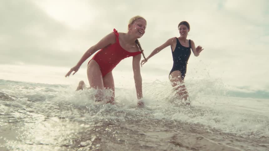 A smiling mother and daughter playfully splash and run through the shallow ocean water in slow motion
