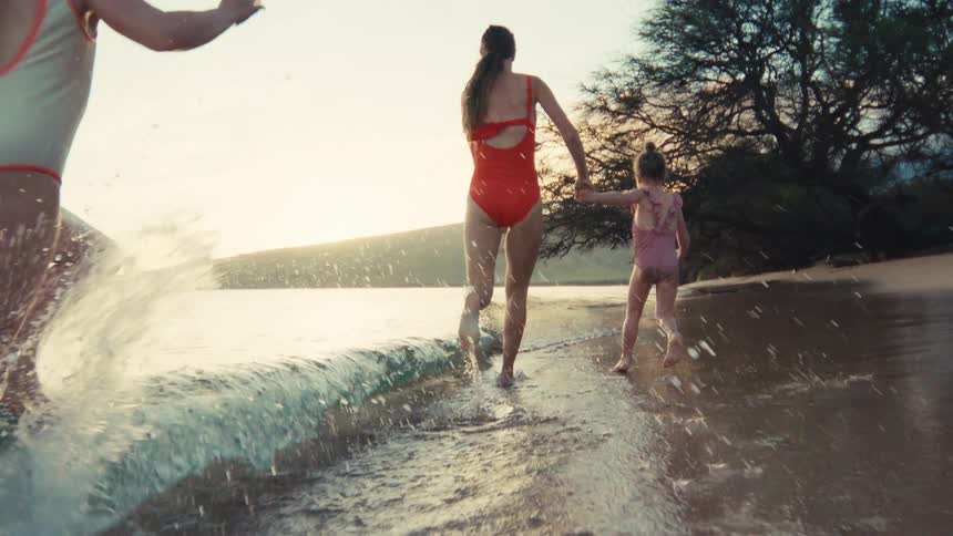 A mother and her two young daughters run hand-in-hand along a beautiful beach at sunset, splashing in the ocean waves in slow motion