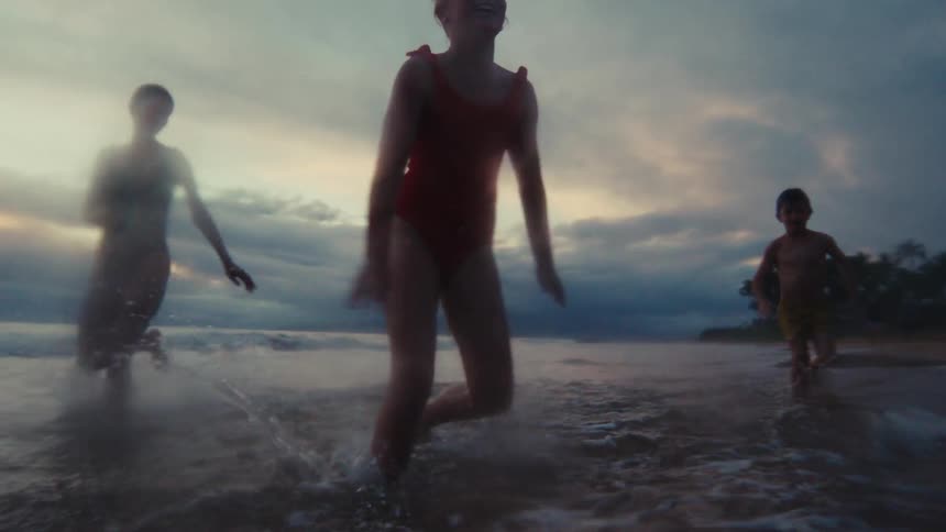 A family playfully runs through the shallow ocean water, splashing and jumping in dreamy slow motion against a dramatic twilight sky