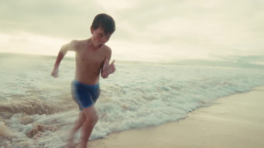 A happy young boy runs along the shoreline in slow motion as the ocean waves crash on the sandy beach