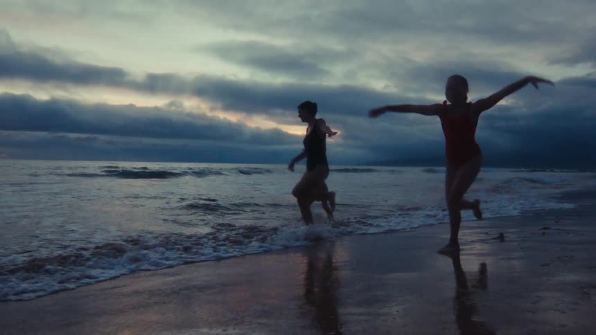 A mother and her children joyfully run and splash in the ocean waves in silhouette against a dramatic twilight sky in slow motion