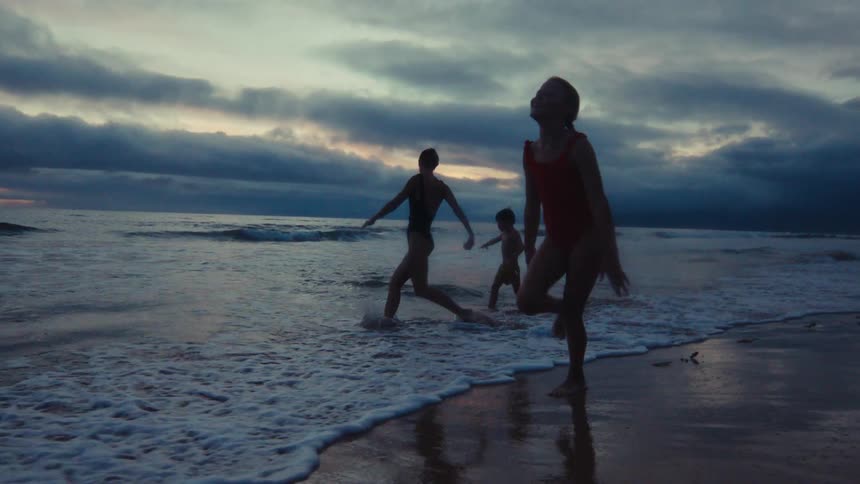 A mother and her children joyfully run and splash in the ocean waves in silhouette against a dramatic twilight sky in slow motion