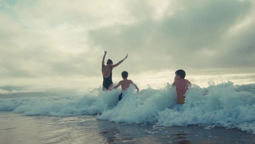A family enjoys a playful day at the beach as they get hit by ocean waves in dramatic slow motion