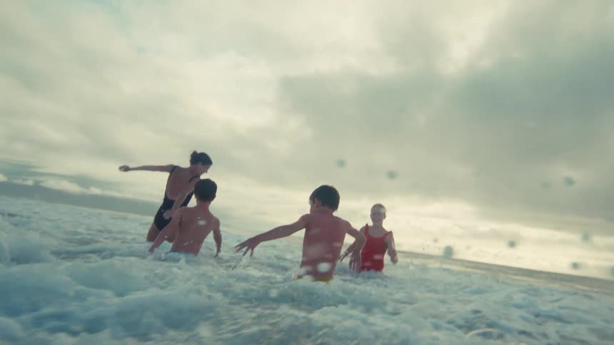 A family enjoys a playful day at the beach as they get hit by ocean waves in dramatic slow motion