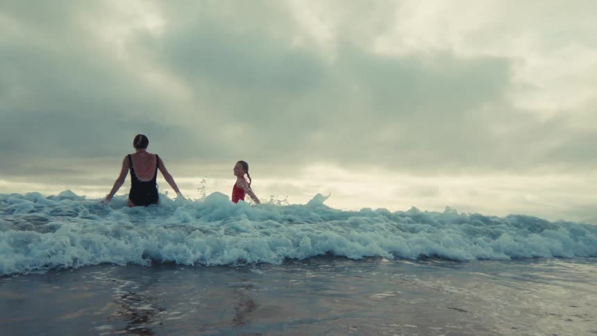 From a low-angle perspective, a powerful ocean wave crashes over two women and the camera in dramatic slow motion under an overcast sky