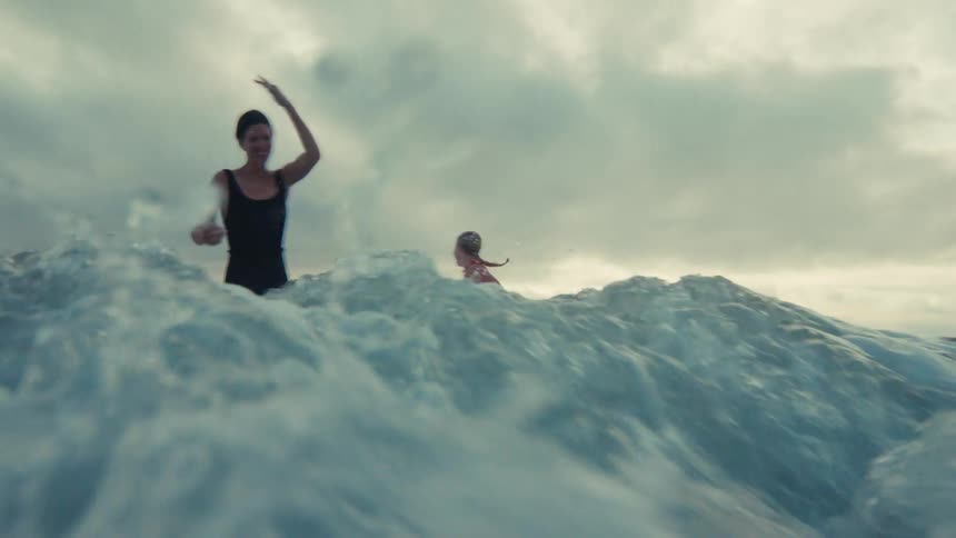 From a low-angle perspective, a powerful ocean wave crashes over two women and the camera in dramatic slow motion under an overcast sky