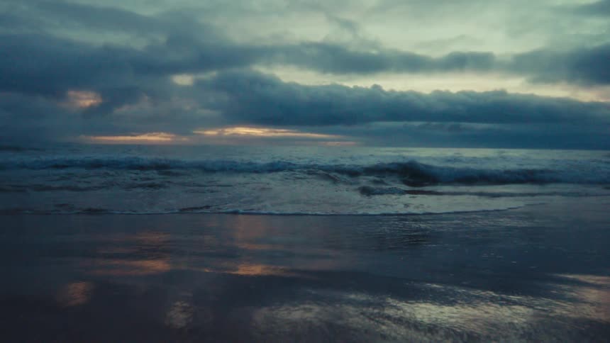 Moody slow motion shot of ocean waves rolling onto the shore under a dramatic cloudy sky at dusk
