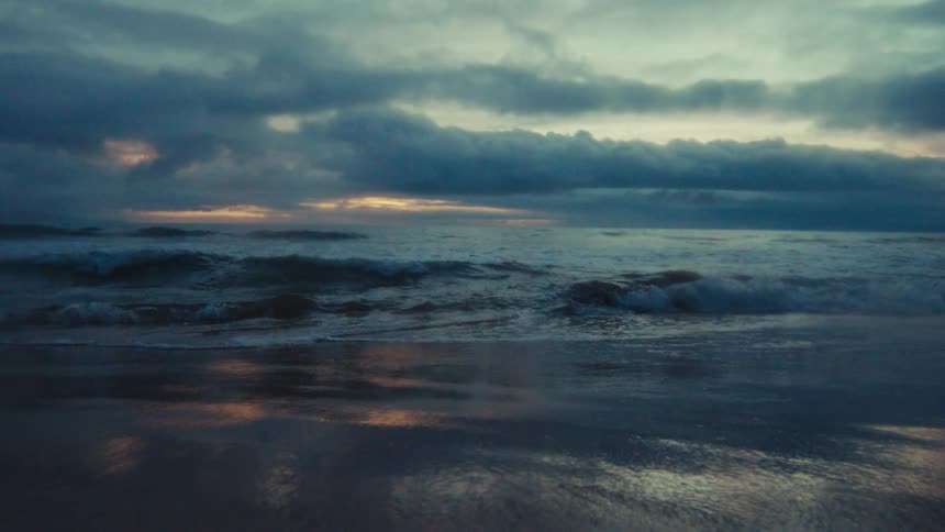 Moody slow motion shot of ocean waves rolling onto the shore under a dramatic cloudy sky at dusk