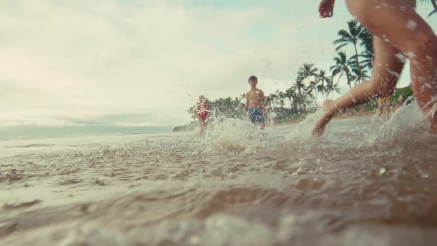 A family in swimsuits happily runs and splashes through the shallow water of a tropical beach in slow motion