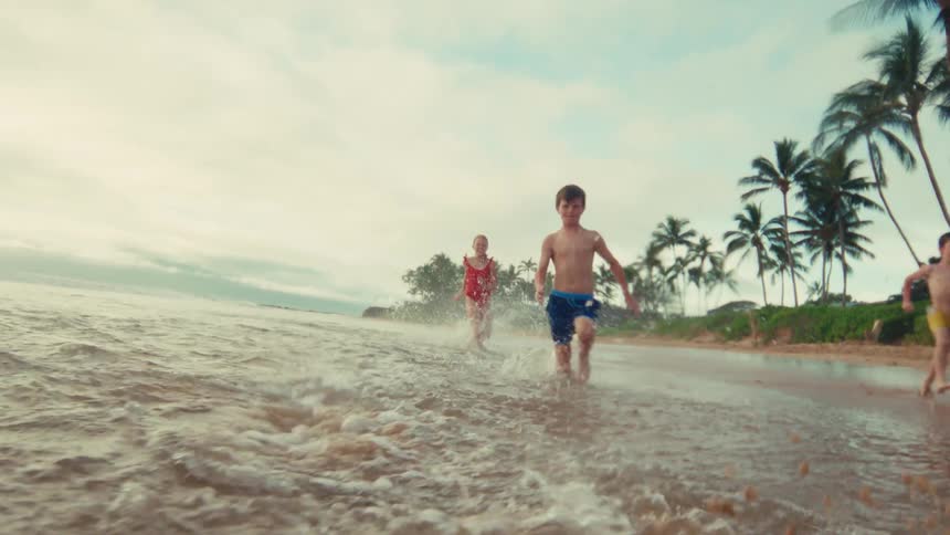 A family in swimsuits happily runs and splashes through the shallow water of a tropical beach in slow motion