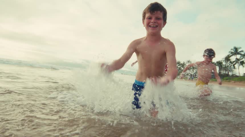 A joyous young boy laughs as he splashes water at the camera while playing with his siblings and mother in the shallow ocean waves in slow motion