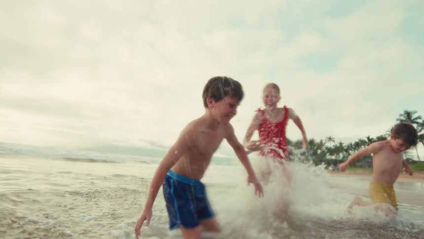 A joyous young boy laughs as he splashes water at the camera while playing with his siblings and mother in the shallow ocean waves in slow motion