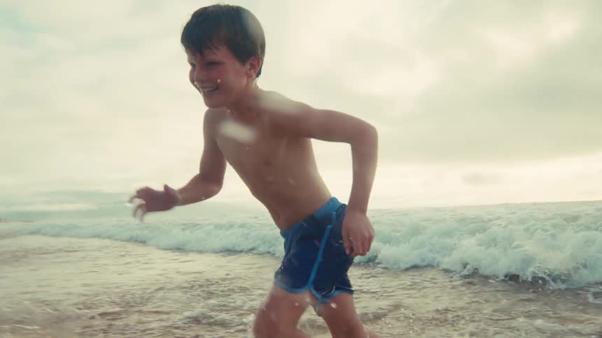 A joyful mother and her young son laugh and run together through the ocean waves in slow motion