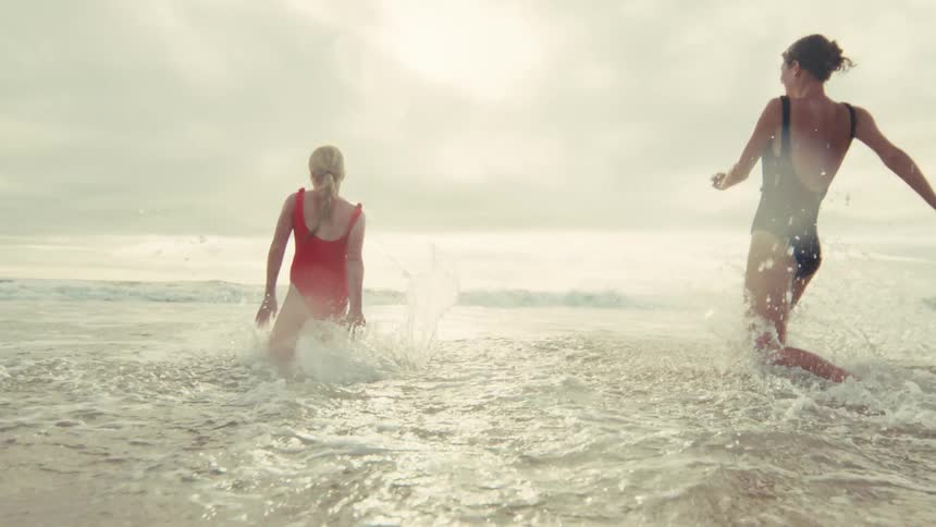 Two joyful young women in swimsuits run and splash into the ocean waves on a tropical beach in slow motion