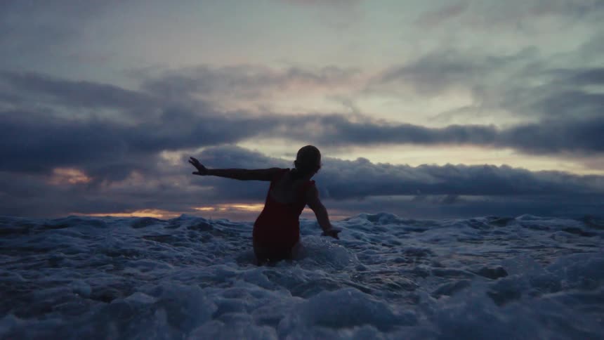 A young girl silhouetted in a red swimsuit joyfully jumps in the ocean waves during a beautiful, cloudy sunset