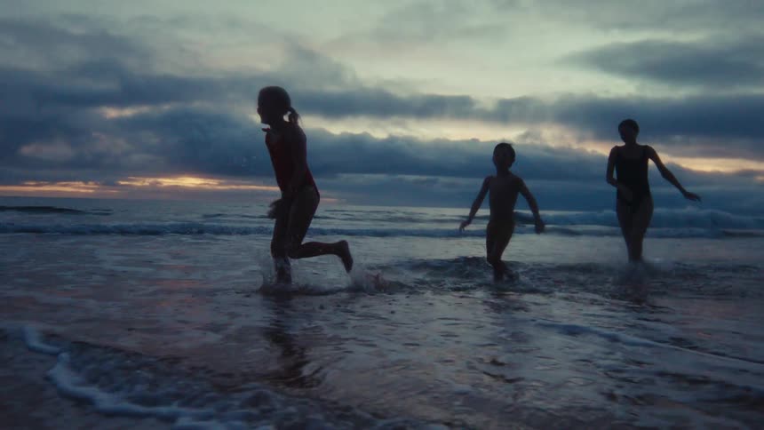 A silhouetted mother and her two children joyfully run and play in the shallow ocean waves at sunset in slow motion