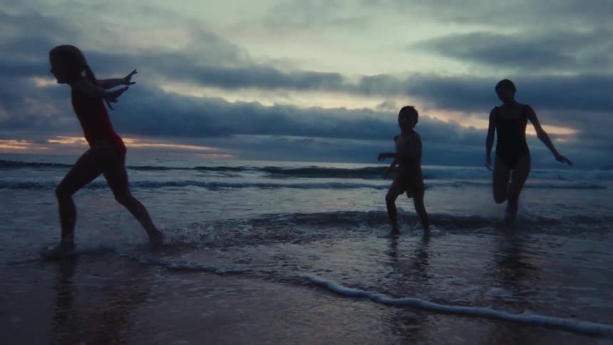 A silhouetted mother and her two children joyfully run and play in the shallow ocean waves at sunset in slow motion