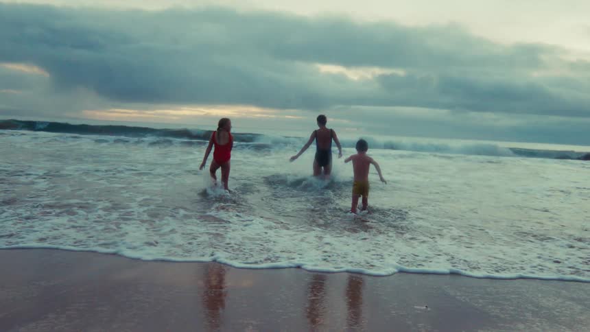 A woman and her two children playfully enter the ocean waves in slow motion under a dramatic, cloudy sky at dusk
