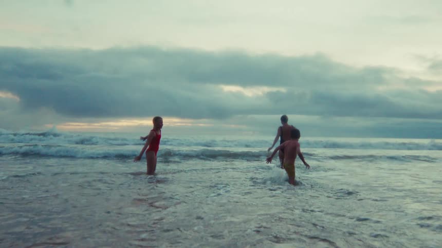 A woman and her two children playfully enter the ocean waves in slow motion under a dramatic, cloudy sky at dusk