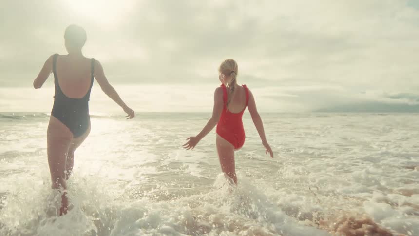 A mother and daughter joyfully run into the ocean waves at a sunny beach in slow motion