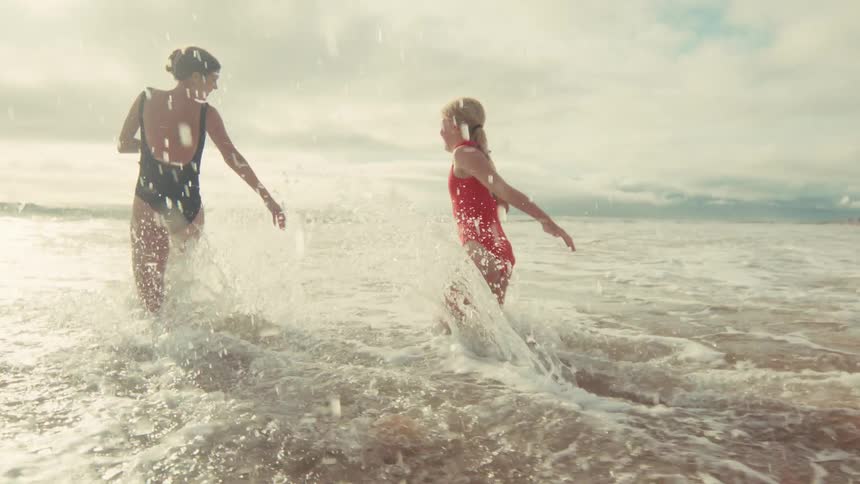 A mother and daughter joyfully run into the ocean waves at a sunny beach in slow motion