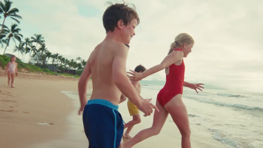 A joyful family with young children excitedly run along the sandy shoreline in slow motion on a beautiful tropical beach