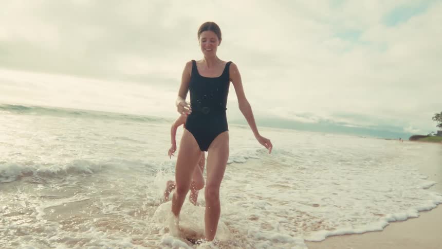 A happy mother and her young daughter run and splash in the gentle ocean waves along a sandy tropical beach in slow motion