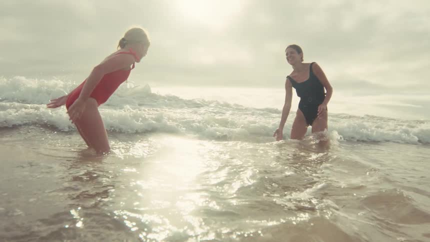 A mother and her young daughter playfully splash and jump in the sunlit ocean waves in slow motion