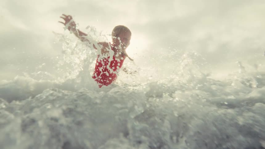 A mother and her young daughter playfully splash and jump in the sunlit ocean waves in slow motion