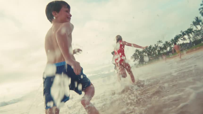 A group of happy kids run and splash in the shallow ocean waves on a tropical beach in slow motion