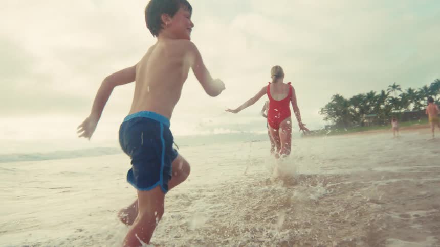 A group of happy kids run and splash in the shallow ocean waves on a tropical beach in slow motion