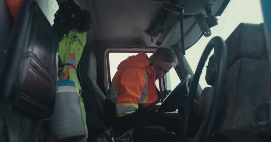 A truck driver in a high-visibility jacket gets into his cab, fastens his seatbelt, and prepares to start his journey