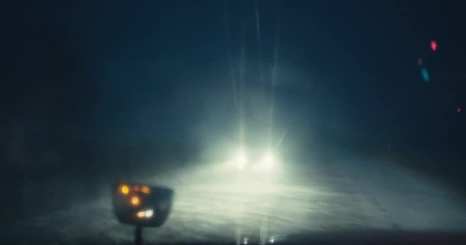 An atmospheric, out-of-focus POV shot from a car driving on a dark, foggy road as an oncoming vehicle's bright lights approach and pass by