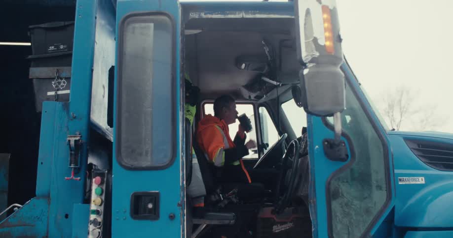 A garbage truck driver in a high-visibility jacket takes a coffee break inside the cab of his truck