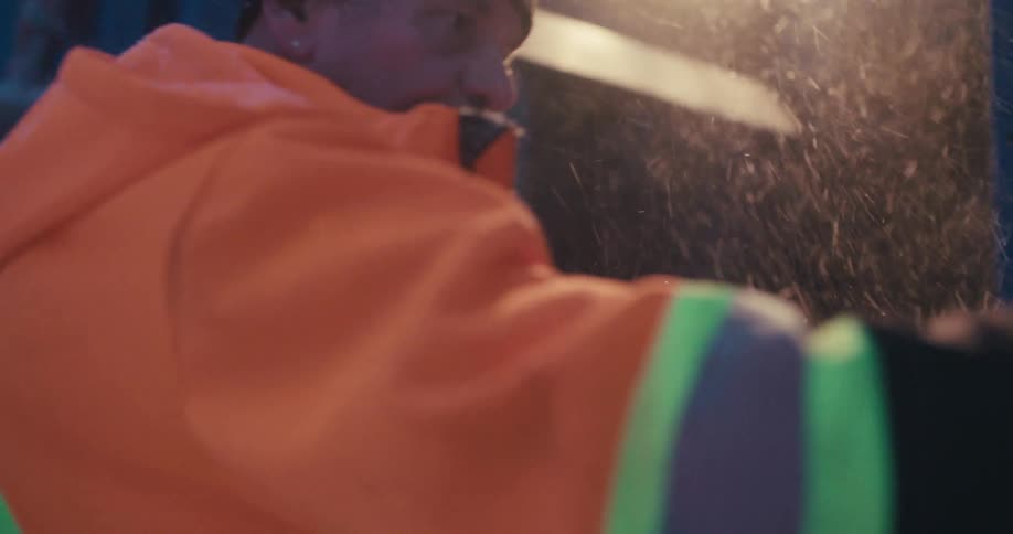 A sanitation worker in an orange high-visibility jacket works on his garbage truck during a cold and snowy night