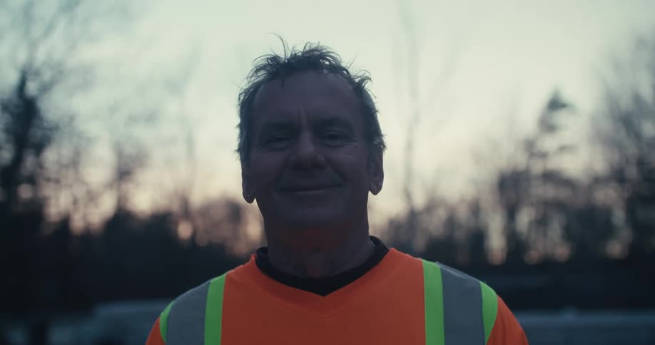 Portrait of a mature blue-collar worker in a safety vest who breaks into a warm smile at dusk