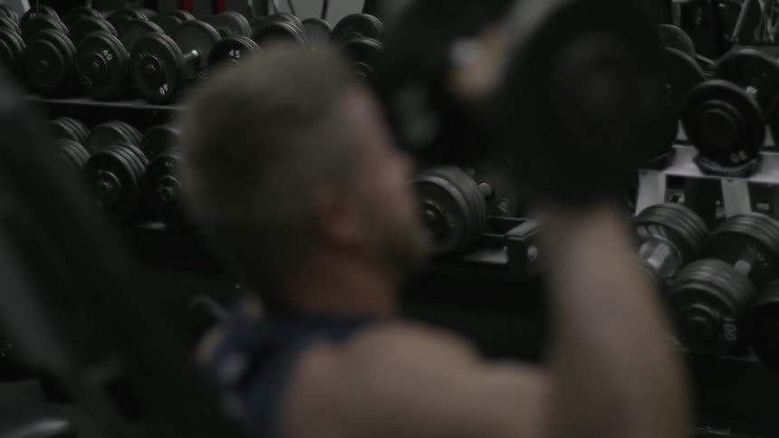 Man sitting on workout bench lifting weight with right arm in gym