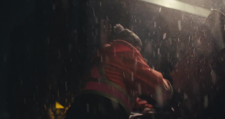 A worker in a beanie and orange safety jacket works relentlessly through a heavy, backlit snowstorm at night