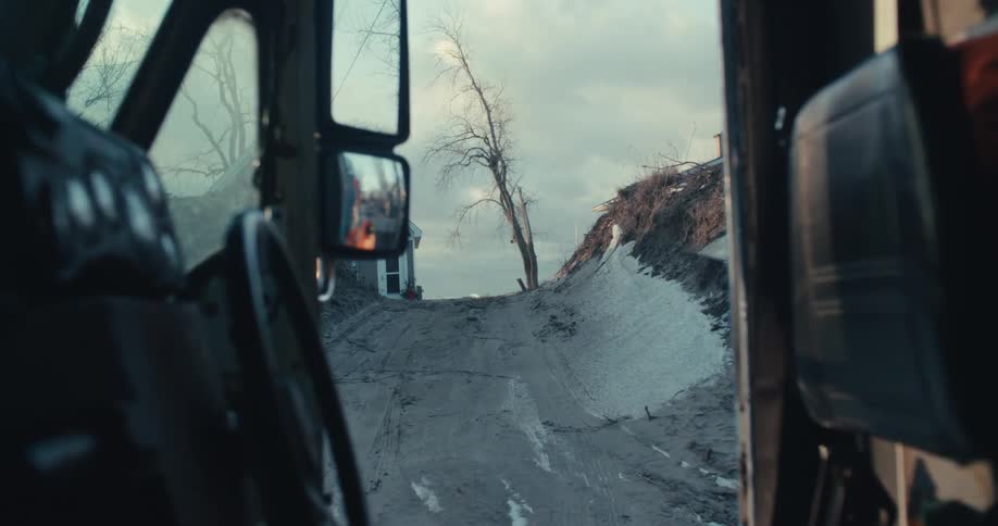 A construction worker in a bright orange jacket gets into the cab of a large truck on a muddy work site
