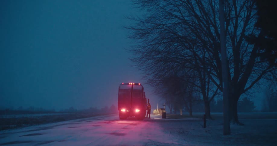 A sanitation worker collects trash on a snowy rural road at dawn, illuminated by the flashing lights of the garbage truck