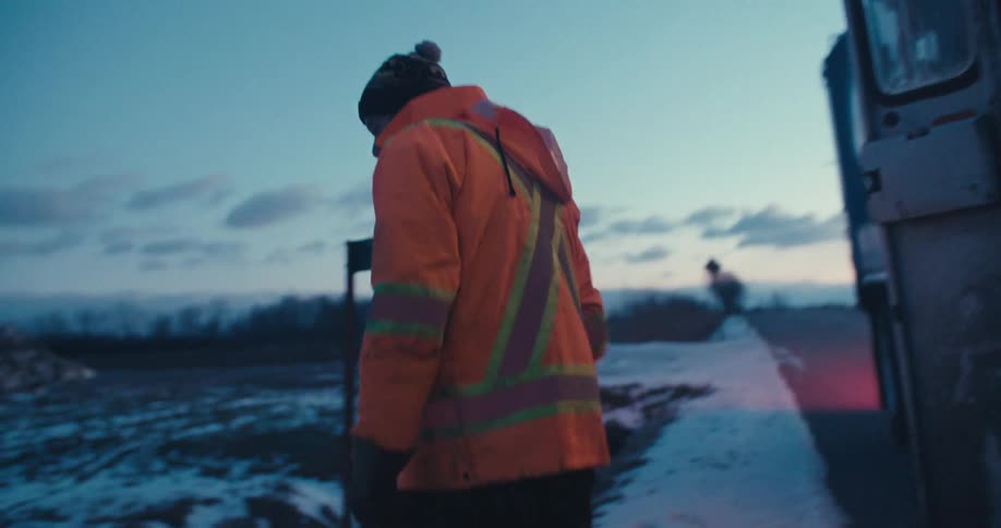 A sanitation worker in high-visibility gear collects a recycling bin from the side of a snowy rural road at dawn