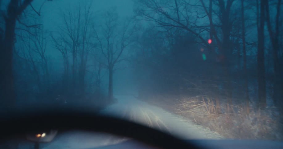 A spooky point-of-view shot from a car driving slowly down a foggy, snow-covered road at night