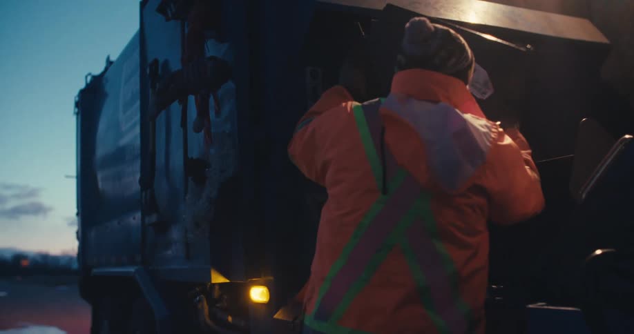 A sanitation worker in an orange safety jacket throws a bag of trash into a garbage truck in the early morning hours
