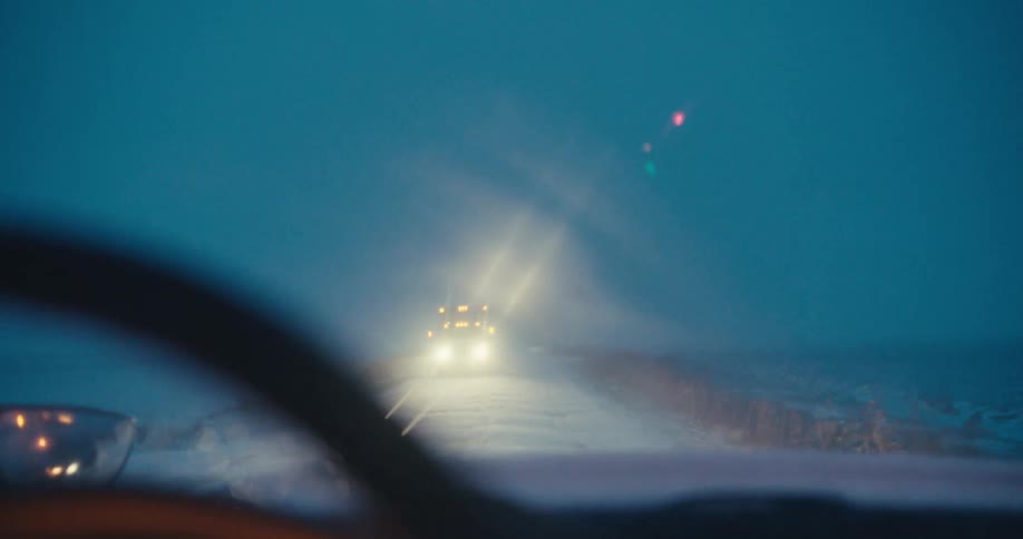 POV shot from inside a car of a large truck approaching with bright headlights on a dark, foggy, and snowy rural road