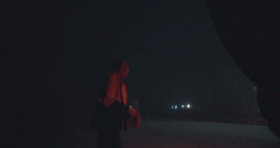 A man in a winter jacket stands on a dark road at night watching the heavy snow fall in slow motion